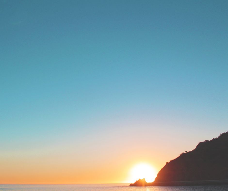 Am Meer geht die Sonne bei blauem Himmel hinter einem Berg auf und strahlt Ruhe und Kraft aus