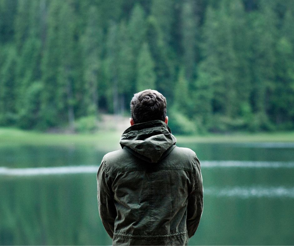 Ein Mann mit braunen Haaren, von hinten zu sehen, blickt in einen ruhigen See. Er trägt einen Parka. Im Hintergrund ein dichter Nadelwald – das Bild strahlt Ruhe und Nachdenklichkeit aus.