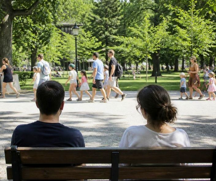 Paar sitzt auf einer Parkbank und blickt auf vorbeigehende Familien mit Kindern.